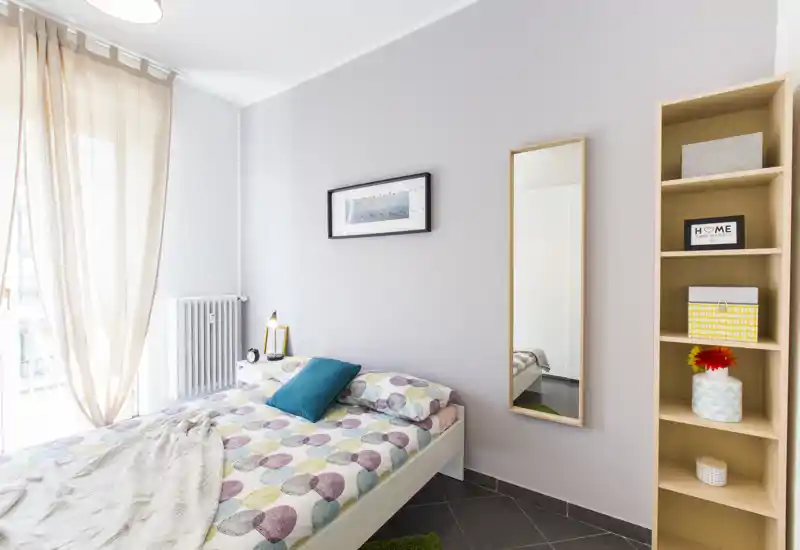 Dormitorio moderno y luminoso con cama doble con ropa de cama estampada, cojín turquesa, espejo de cuerpo entero y estantería; gran ventana con cortinas semitransparentes que aporta mucha luz natural.