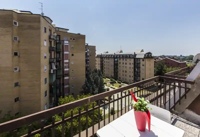 Balcone soleggiato con piccolo tavolo e vaso che guarda edifici residenziali e un cortile con giardini — vista cittadina luminosa, perfetta per il caffè mattutino.