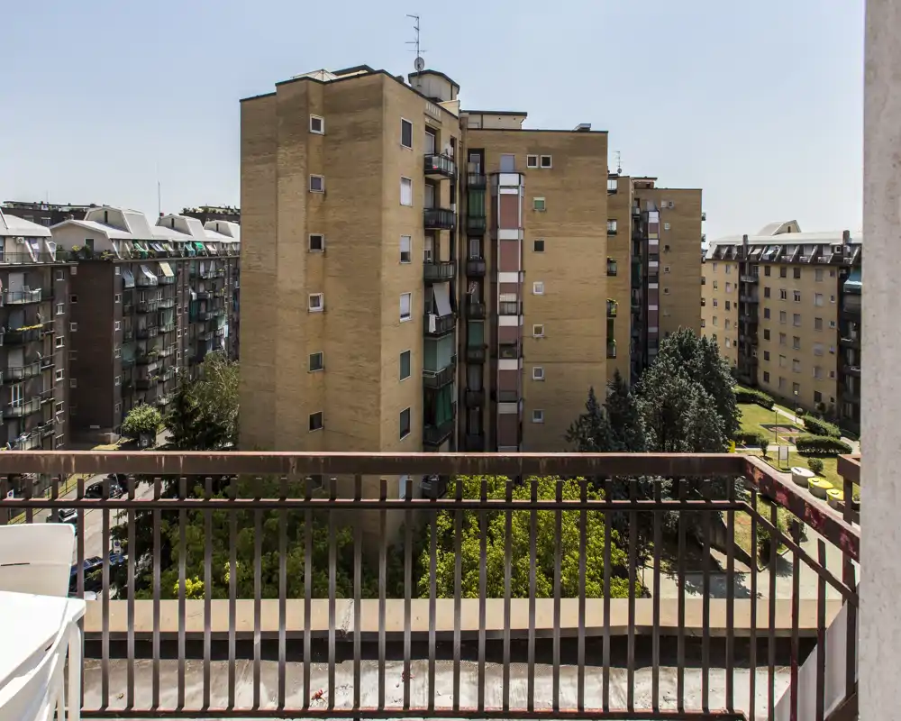 Balcón soleado con vistas a bloques de viviendas y un patio ajardinado. Ideal para tomar un café por la mañana con cielo despejado y zonas verdes.
