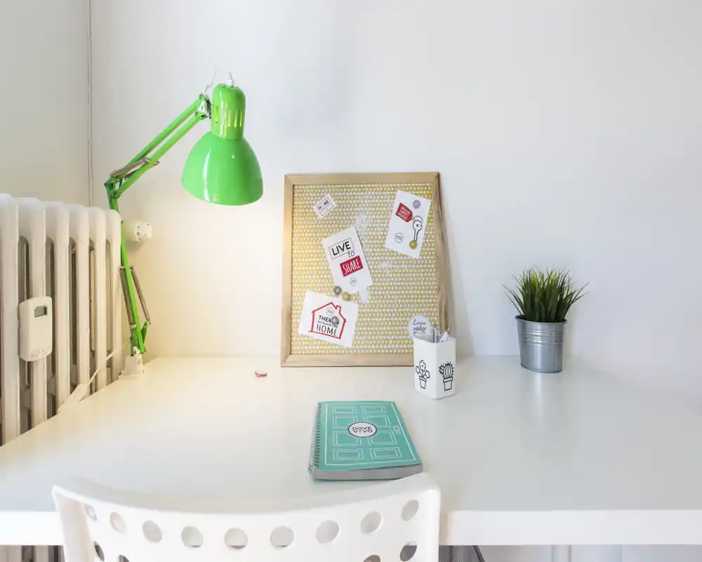 Rincón de escritorio luminoso y minimalista con una lámpara verde articulada, tablero de notas, planta pequeña y silla blanca — perfecto para una zona de estudio o teletrabajo.