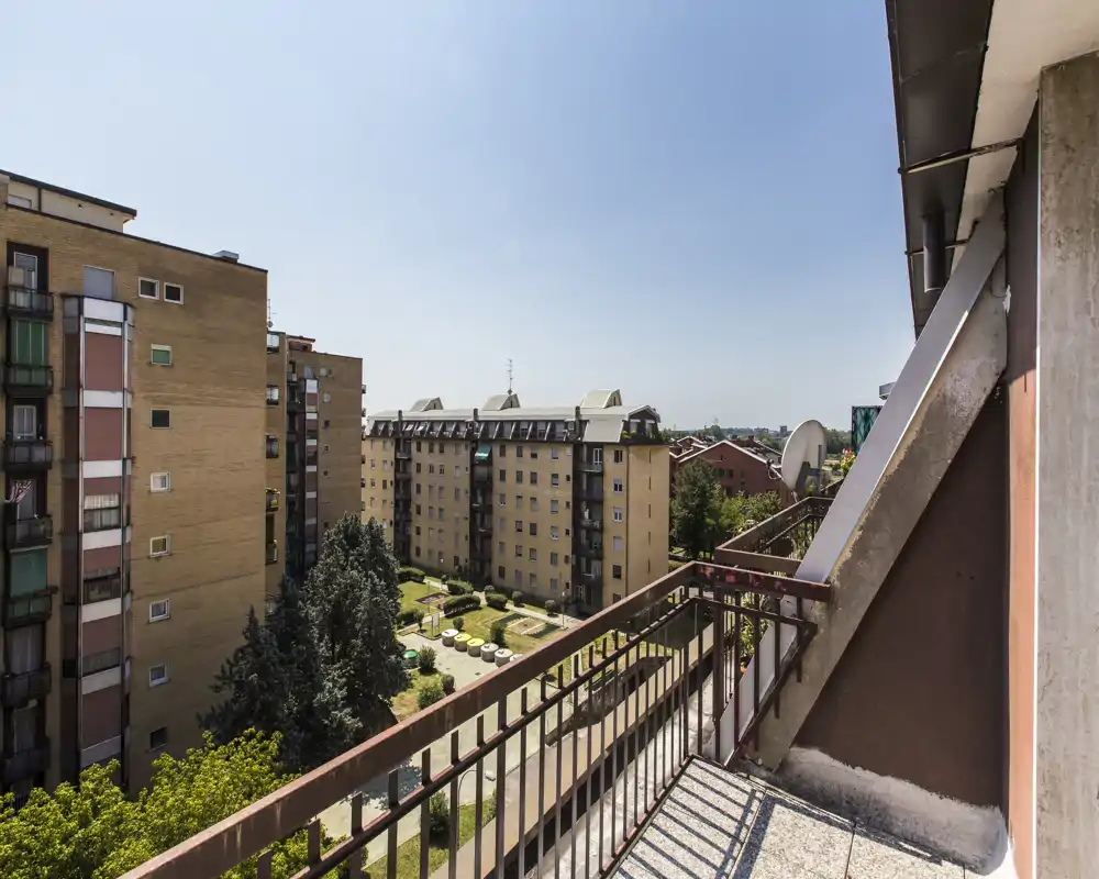 Balcón soleado con barandilla metálica con vistas a un patio ajardinado y edificios residenciales; cielo despejado y agradable vista urbana.
