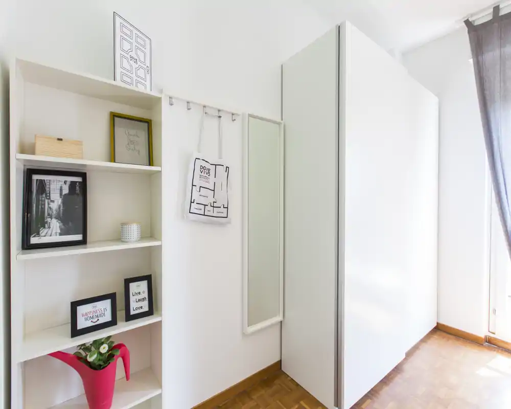 Entrada luminosa con un armario blanco alto, espejo de pared y estantería con marcos decorativos y una planta. Luz natural entra por la derecha.