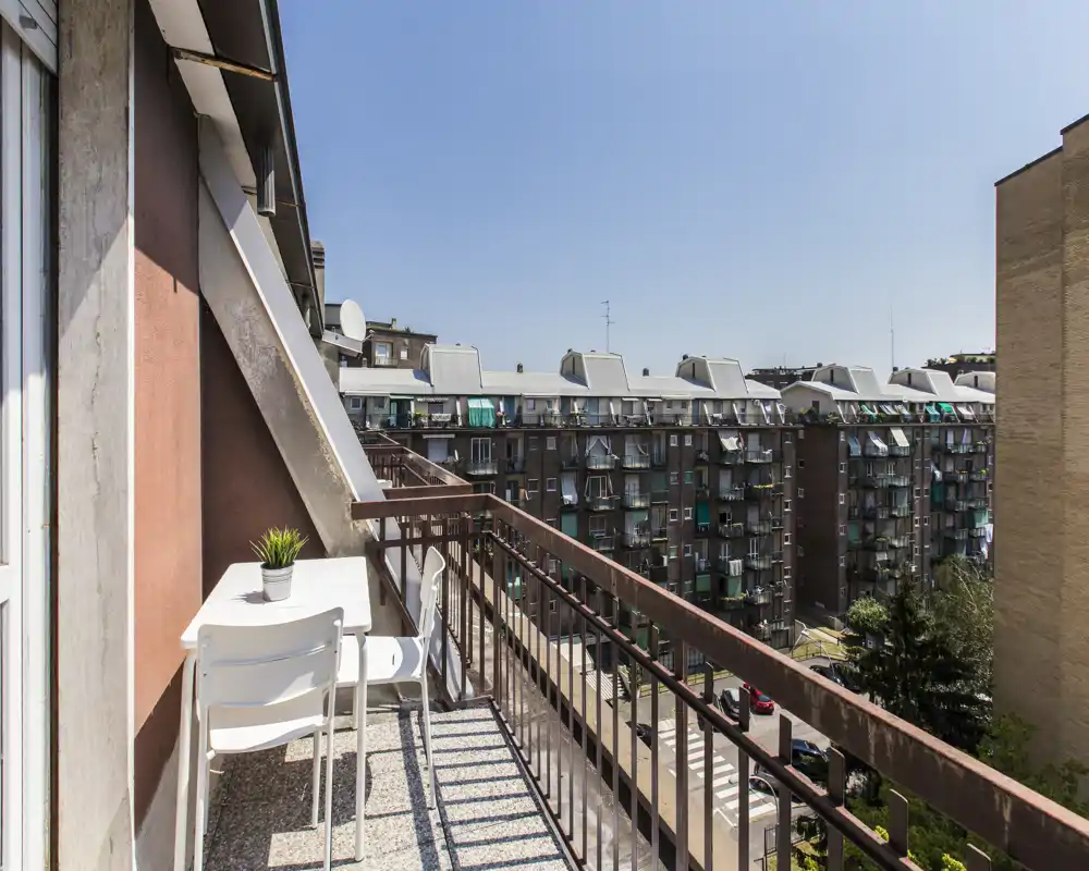 Balcón privado soleado con una pequeña mesa blanca y dos sillas, planta en maceta y vistas despejadas a los edificios vecinos — ideal para el café de la mañana.