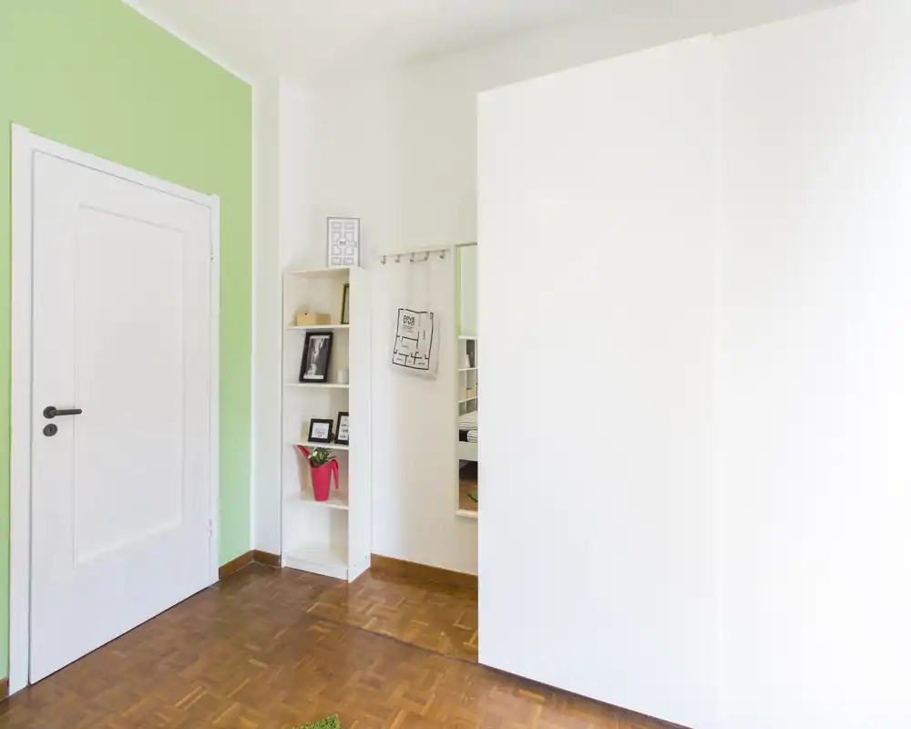 Rincón de dormitorio luminoso y minimalista con armario blanco, estantería y un reflejo parcial en el espejo que sugiere la cama fuera del encuadre. Parquet y pared verde ofrecen sensación limpia y aireada.