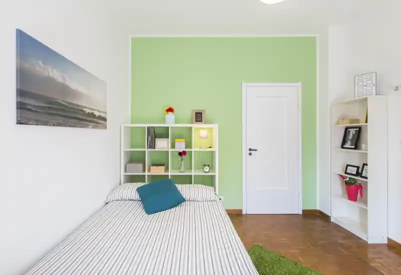 Dormitorio individual moderno y luminoso con cama a rayas, cojín turquesa, estanterías abiertas y pared de acento verde claro — sencillo y bien iluminado.