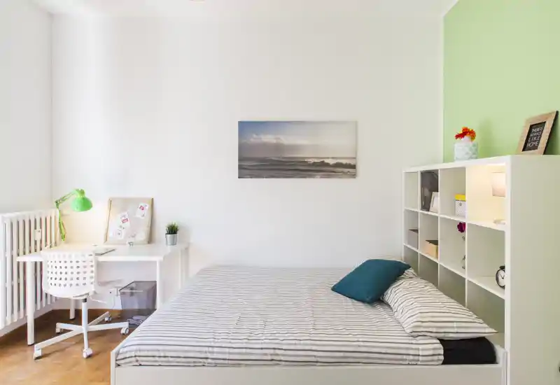 Dormitorio luminoso y moderno con cama doble, ropa de cama a rayas y un cojín turquesa. Un escritorio compacto con lámpara verde está junto al radiador y una estantería de cubos blanca ofrece almacenamiento y decoración.