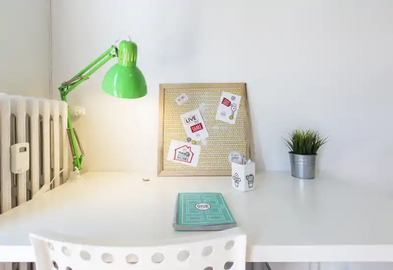 Coin bureau lumineux et minimaliste avec lampe de bureau verte réglable, tableau d'affichage, petite plante et chaise blanche — idéal pour un espace de travail à domicile.