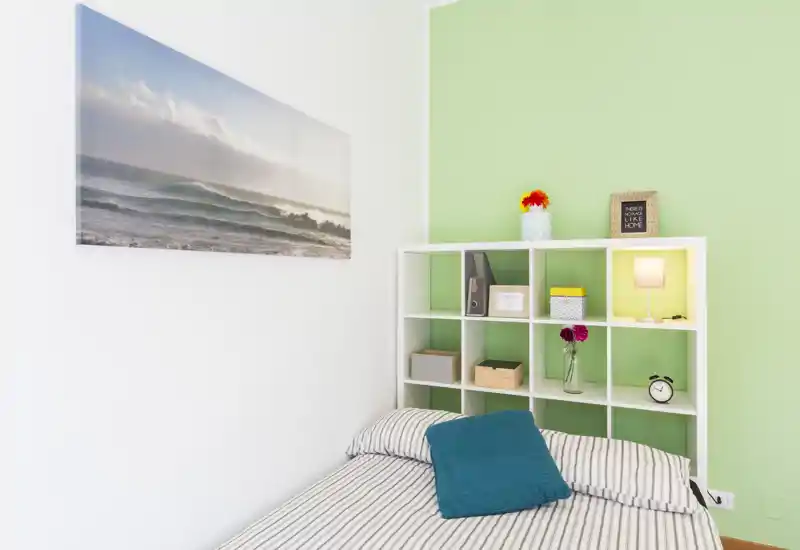 Rincón de dormitorio luminoso y ordenado con cama de rayas, cojín azul verdoso y estantería abierta como cabecero; iluminación suave y cuadro costero que crean un ambiente fresco y acogedor.