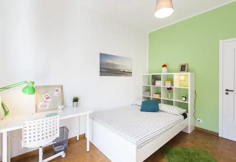 Dormitorio luminoso y moderno con cama doble, estantería como cabecero, escritorio y pared verde de acento — ideal para una estancia acogedora.