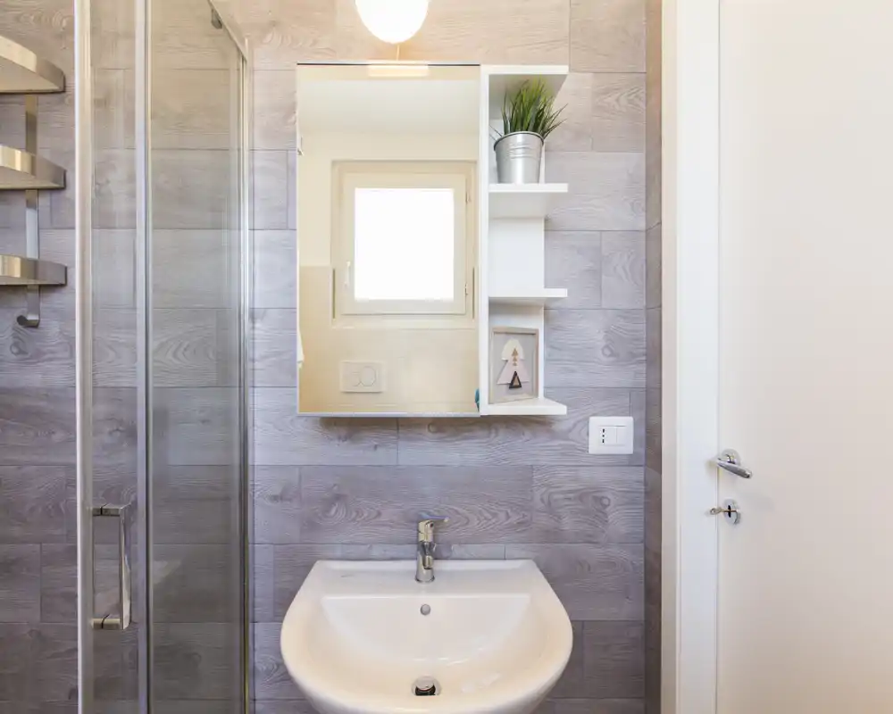 Baño moderno y luminoso con lavabo blanco, armario con espejo y puerta de ducha de cristal. Azulejos efecto madera y una balda decorativa crean un espacio limpio y acogedor.