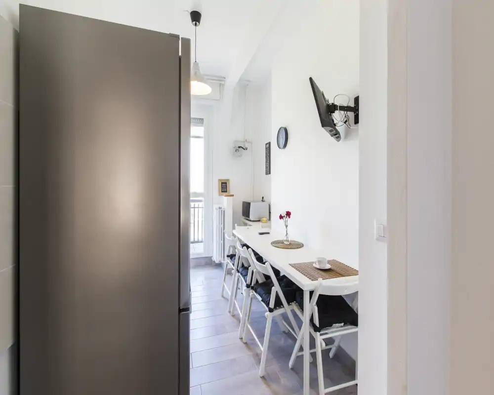 Cocina/comedor estrecho y moderno con mesa alargada, sillas plegables y luz natural desde la puerta al balcón — ordenado, luminoso y funcional para comer.