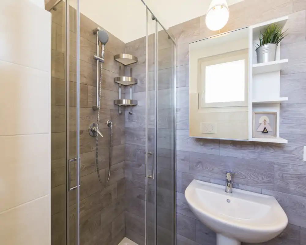 Baño moderno y compacto con mampara de ducha de vidrio, grifería de pared y lavabo blanco sobre pedestal bajo un mueble con espejo.