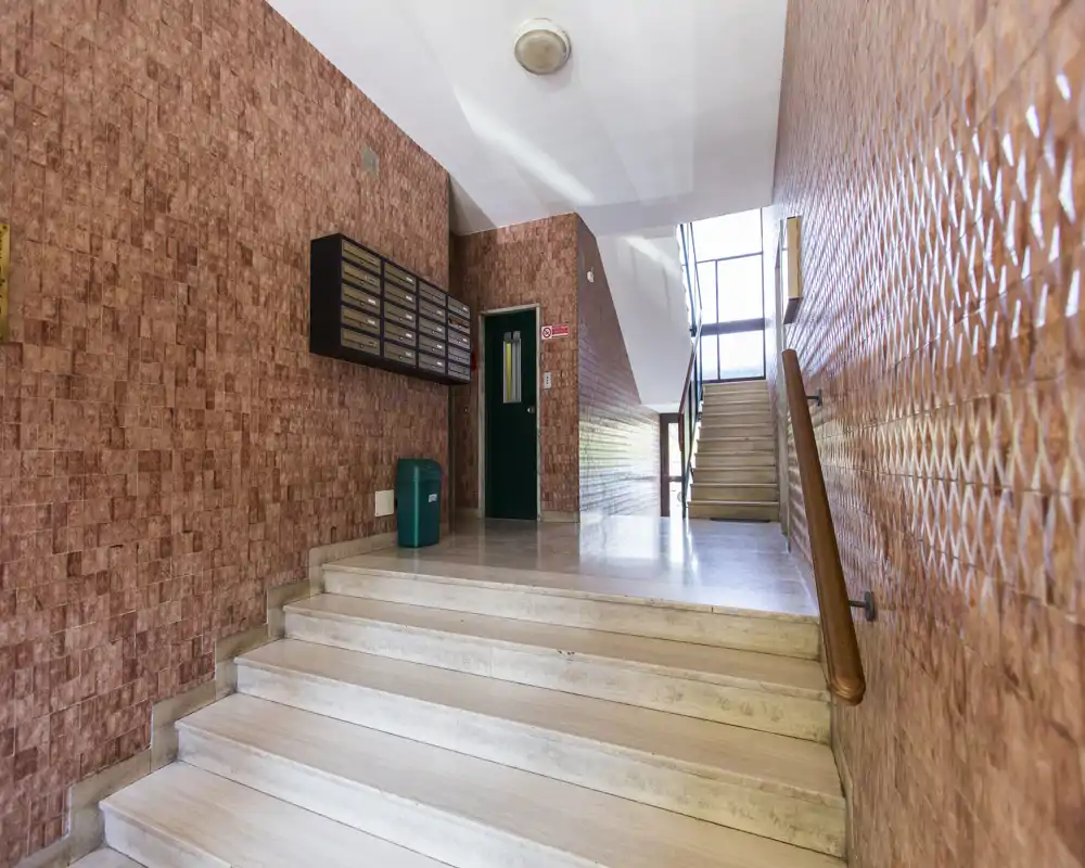 Vestíbulo de edificio luminoso con escalones de mármol, una escalera que sube, buzones en la pared y pasamanos — zona común bien iluminada y ordenada.
