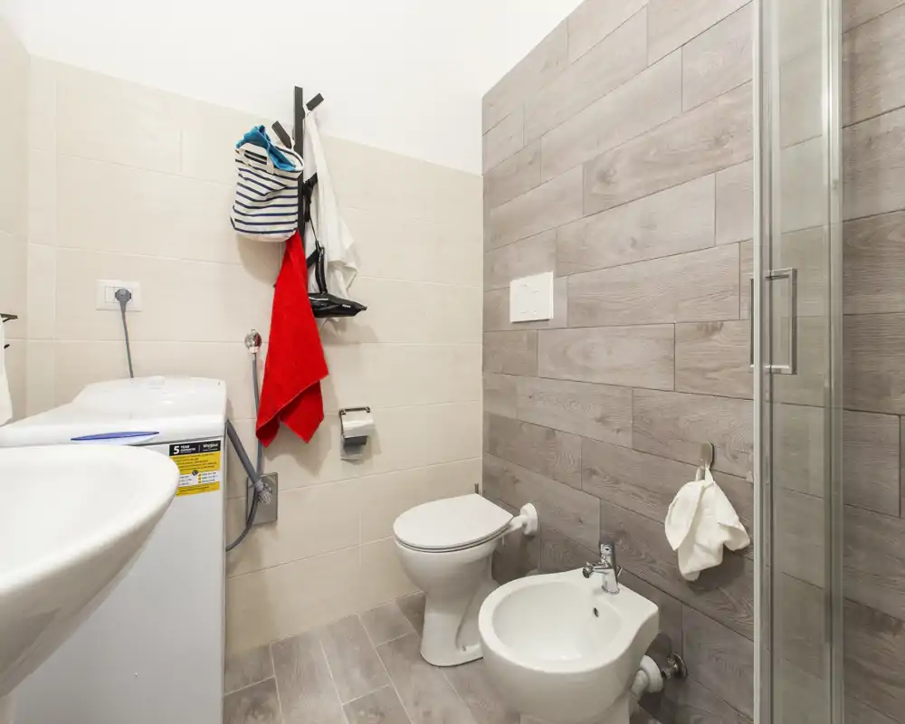 Baño moderno y luminoso con inodoro, bidé, lavabo y ducha en esquina; revestimiento efecto madera y lavadora para mayor comodidad.