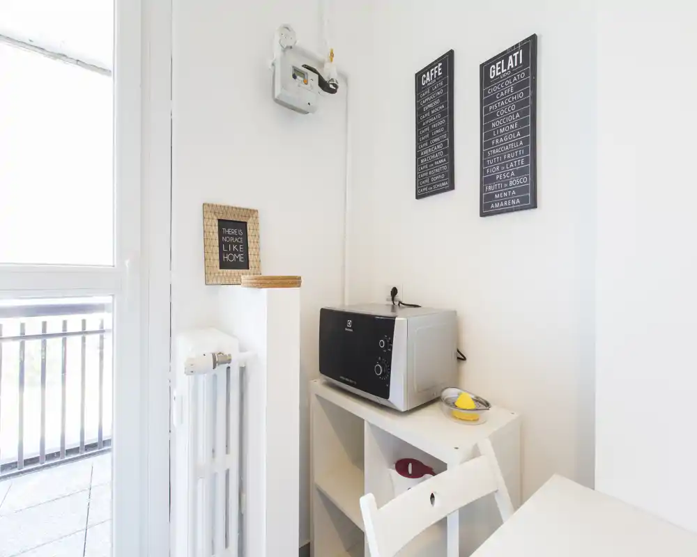 Rincón de kitchenette compacto con microondas sobre una pequeña estantería, una silla blanca y luz natural intensa desde la puerta al balcón.