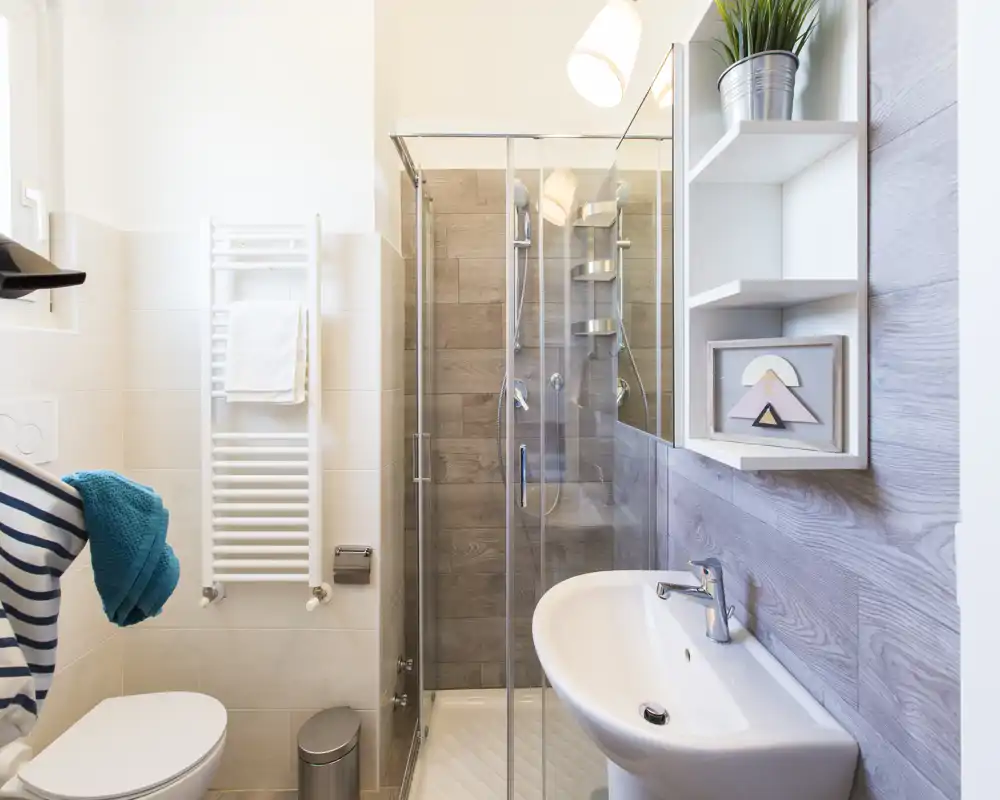 Baño moderno y luminoso con ducha acristalada, lavabo de pared y WC, azulejos efecto madera y estanterías prácticas.