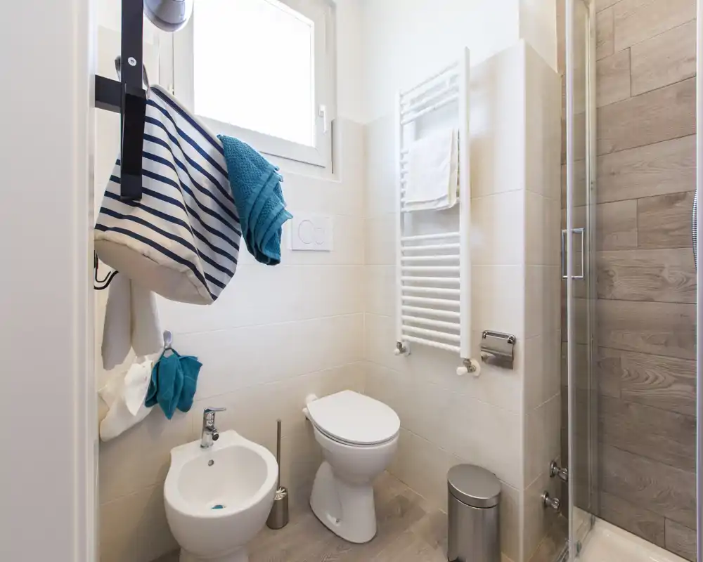 Baño moderno y compacto con inodoro, bidé y ducha con mampara de vidrio. Baldosas neutras y panel efecto madera dan un aspecto limpio y fresco.