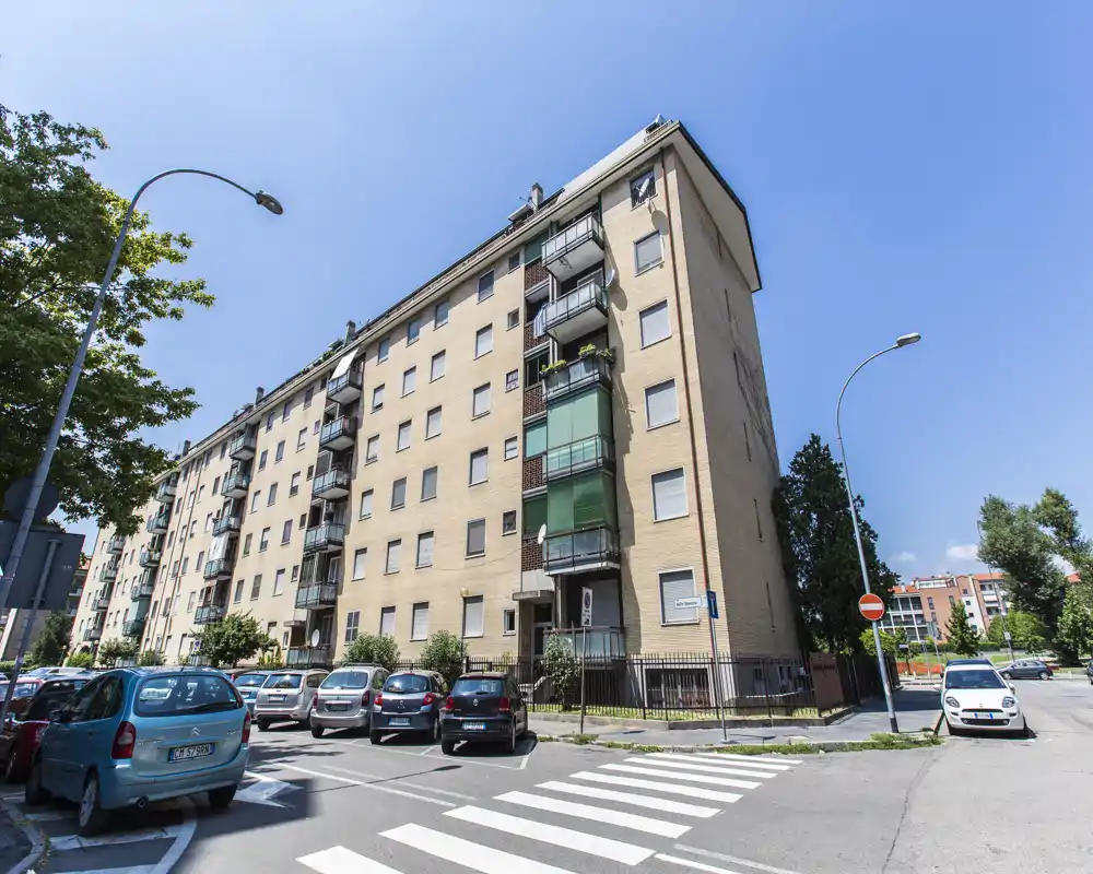 Vista exterior de un edificio de apartamentos de altura media con balcones y coches aparcados, bajo un cielo azul despejado — útil para mostrar la fachada y la calle alrededor.