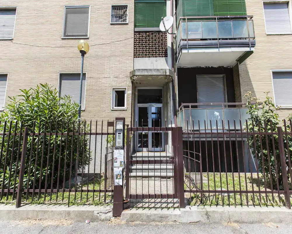 Vista exterior de la entrada de un edificio con reja, escalones hacia la puerta y un balcón en el piso superior.