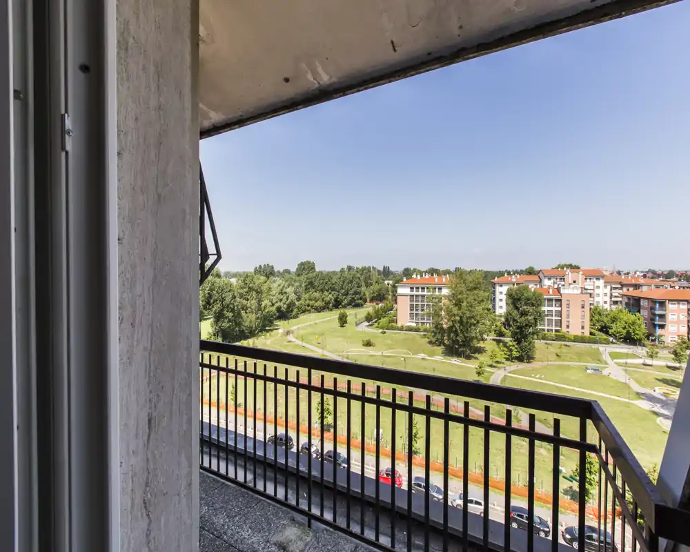 Balcón soleado con barandilla metálica con vistas a un parque verde y edificios residenciales, con una agradable vista abierta ideal para relajarse al aire libre.