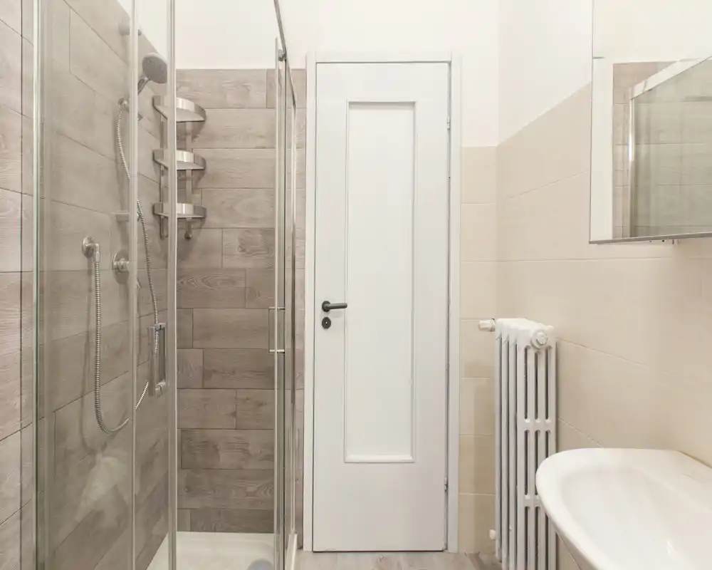 Baño moderno y compacto con ducha acristalada, grifería de pared y lavabo visible a la derecha. Tonos neutros y líneas limpias que dan un espacio funcional y ordenado.