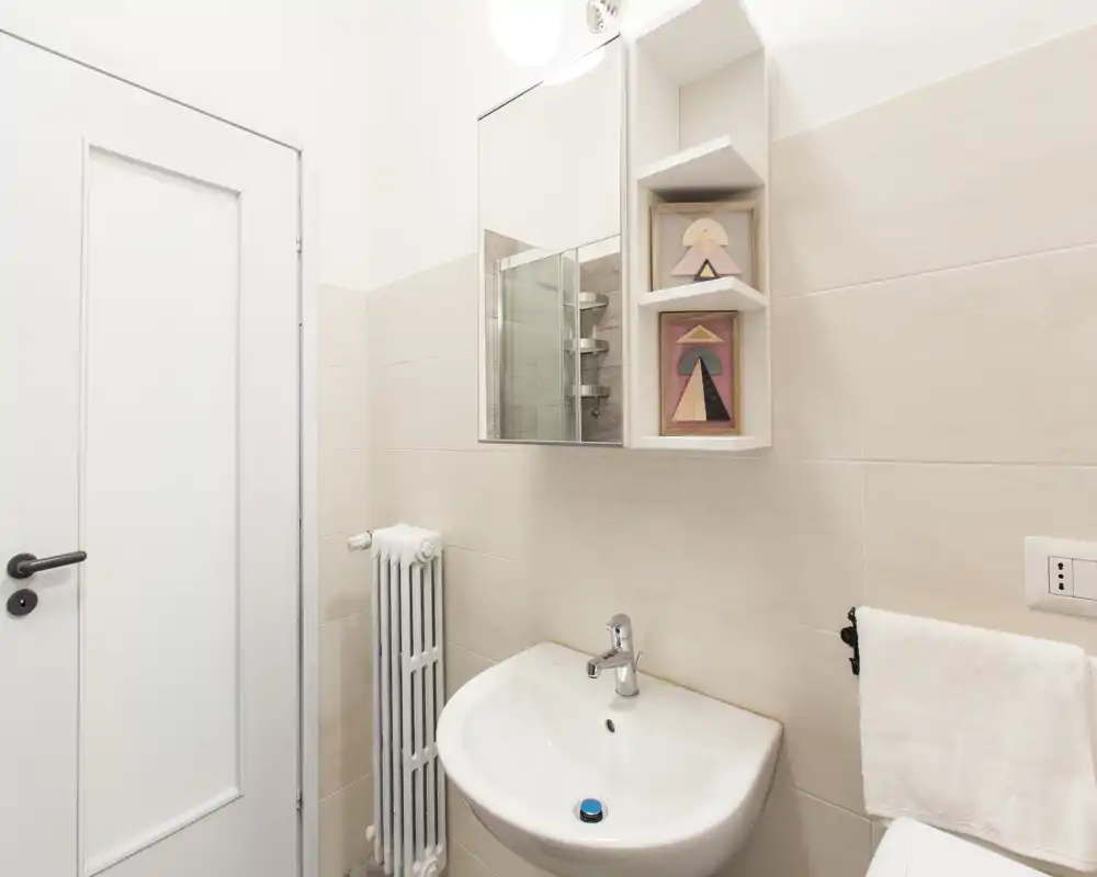 Baño pequeño y moderno, luminoso, con lavabo mural, armario con espejo y toallero. Azulejos beige y sanitarios blancos que transmiten limpieza.