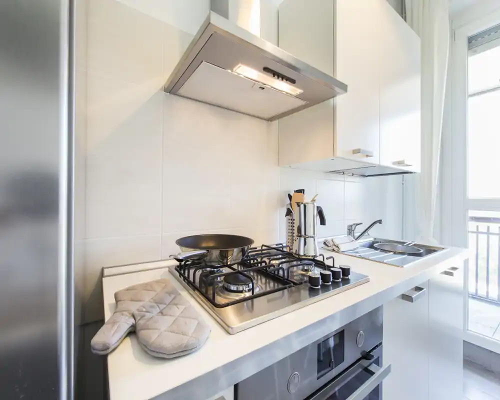 Cocina moderna y compacta con placa de gas de acero inoxidable, campana extractora, fregadero y luz natural; muebles blancos limpios y accesorios prácticos como manopla y utensilios.
