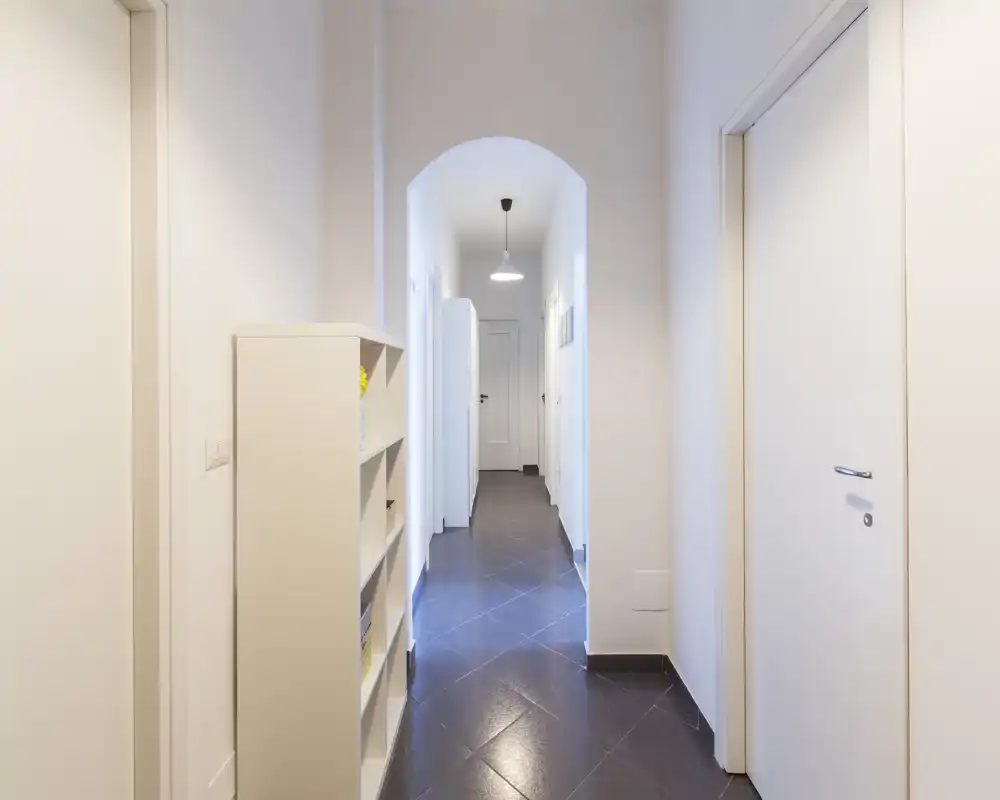 Pasillo luminoso y estrecho con suelo de baldosas, paredes blancas y un arco que conduce a las habitaciones interiores; estantería sencilla y puertas a lo largo del pasillo.