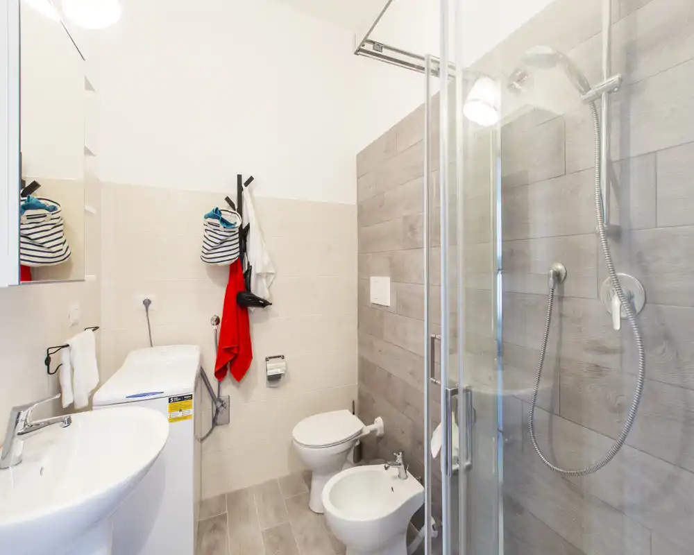 Baño moderno y luminoso con ducha de cristal, inodoro suspendido, bidé y lavabo — azulejos neutros y equipamiento funcional.