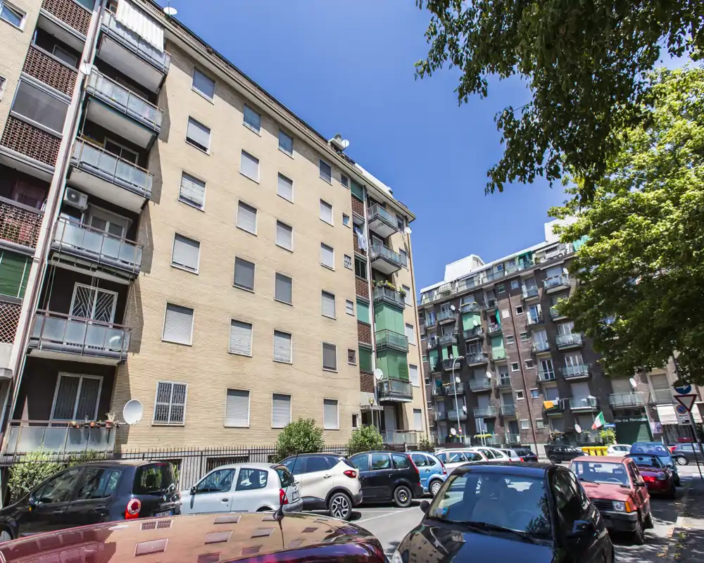 Exterior urbano soleado con edificios de apartamentos, coches aparcados y una calle con árboles — ideal para destacar la ubicación y el aparcamiento.