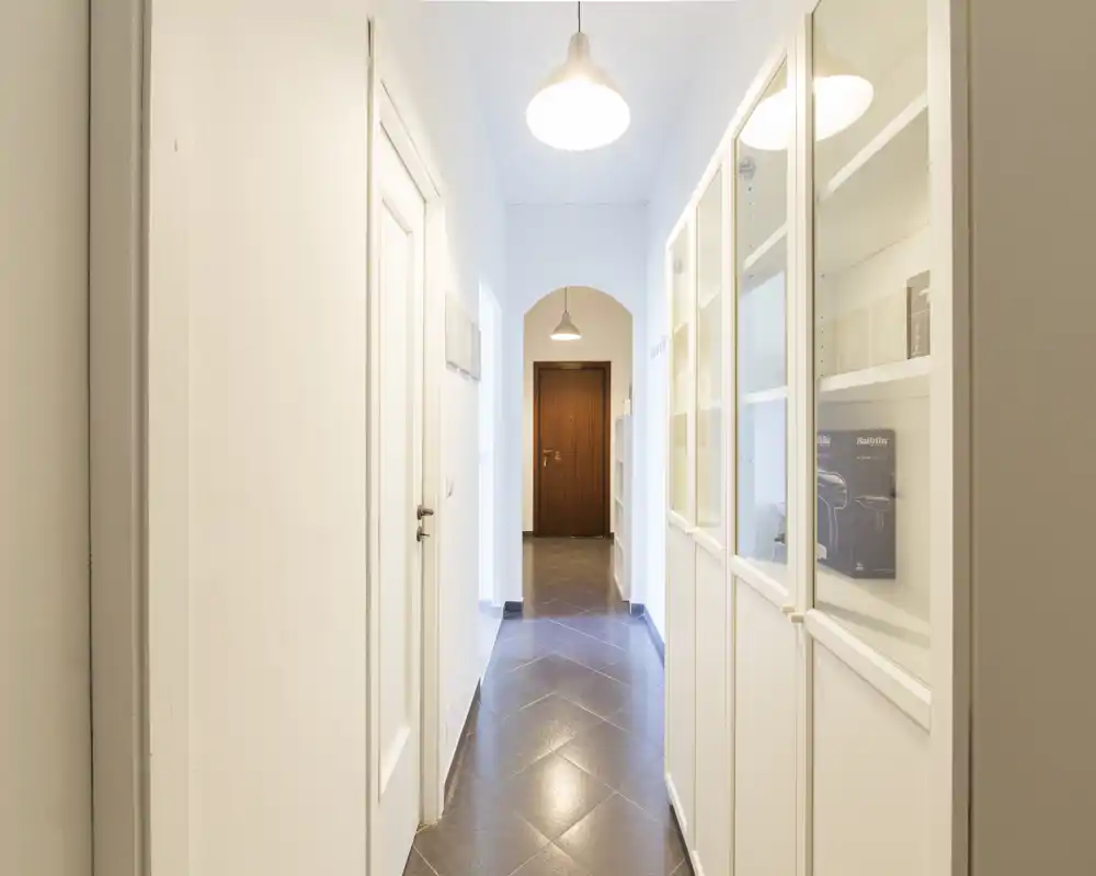 Pasillo de entrada luminoso y estrecho con suelo de baldosas, lámparas colgantes y armarios blancos empotrados — un espacio de entrada limpio y bien iluminado.