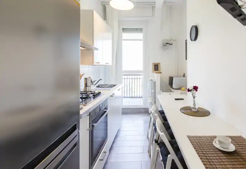 Cuisine en longueur moderne avec meubles blancs, four encastré et plaque à gaz, petit bar avec tabourets et lumière naturelle provenant de la porte du balcon.