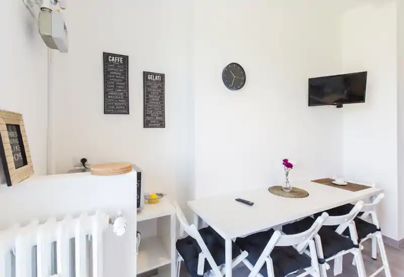 Coin repas moderne et lumineux avec une table blanche, des chaises pliantes rembourrées et une TV murale. Décor épuré avec un petit vase fleuri et des tableaux minimalistes.