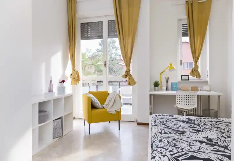 Dormitorio luminoso y moderno con cama estampada, un sillón mostaza y un escritorio junto a la ventana que da a un pequeño balcón: luminoso y bien arreglado para un anuncio.