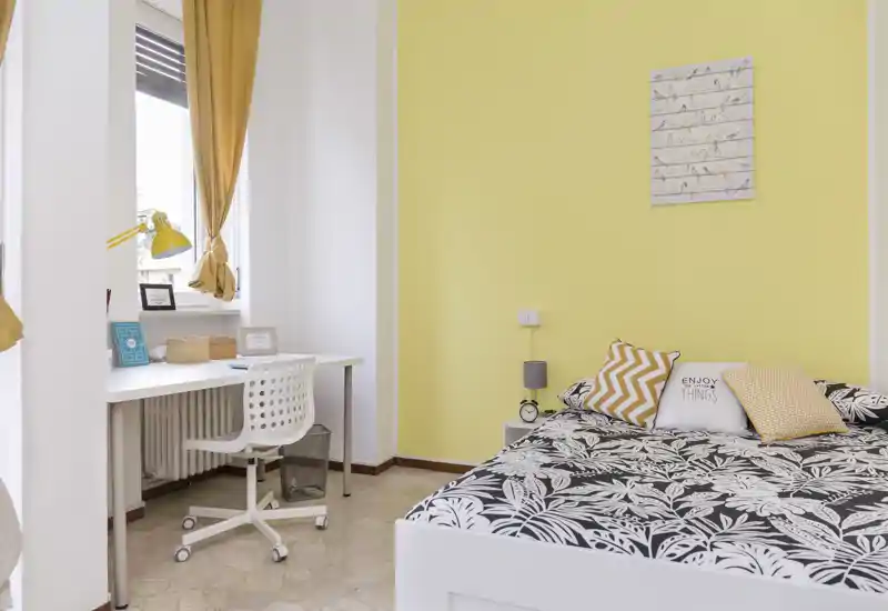 Dormitorio luminoso y acogedor con colcha estampada y pared amarilla; un pequeño escritorio junto a la ventana ofrece un agradable espacio de trabajo.