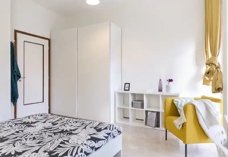 Dormitorio moderno y luminoso con cama con estampado en primer plano, amplio armario blanco y un acogedor sillón amarillo junto a la ventana, ideal para una estancia cómoda.