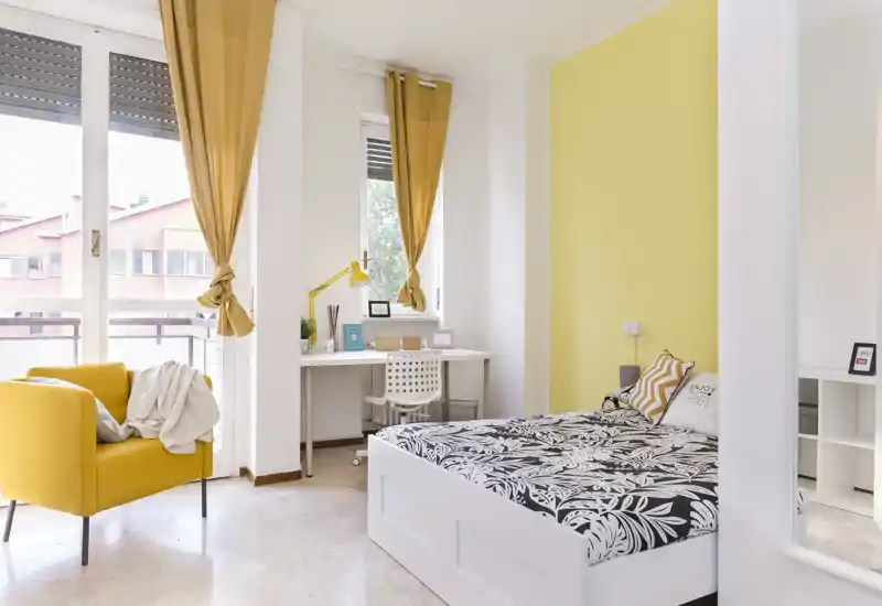 Bright, modern bedroom with a patterned white bedframe and coordinated yellow accents. Large windows flood the room with natural light and a small desk creates a functional workspace.