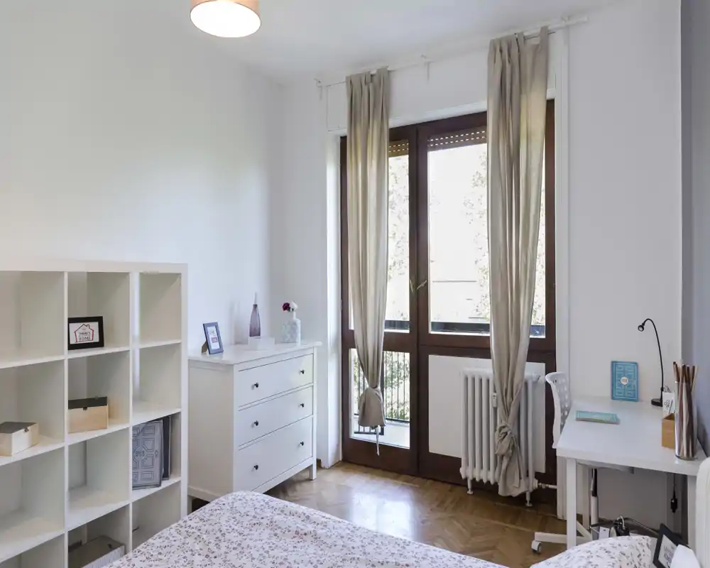 Bright, well-staged bedroom with a patterned bedcover, white storage units and large balcony windows that flood the room with natural light.