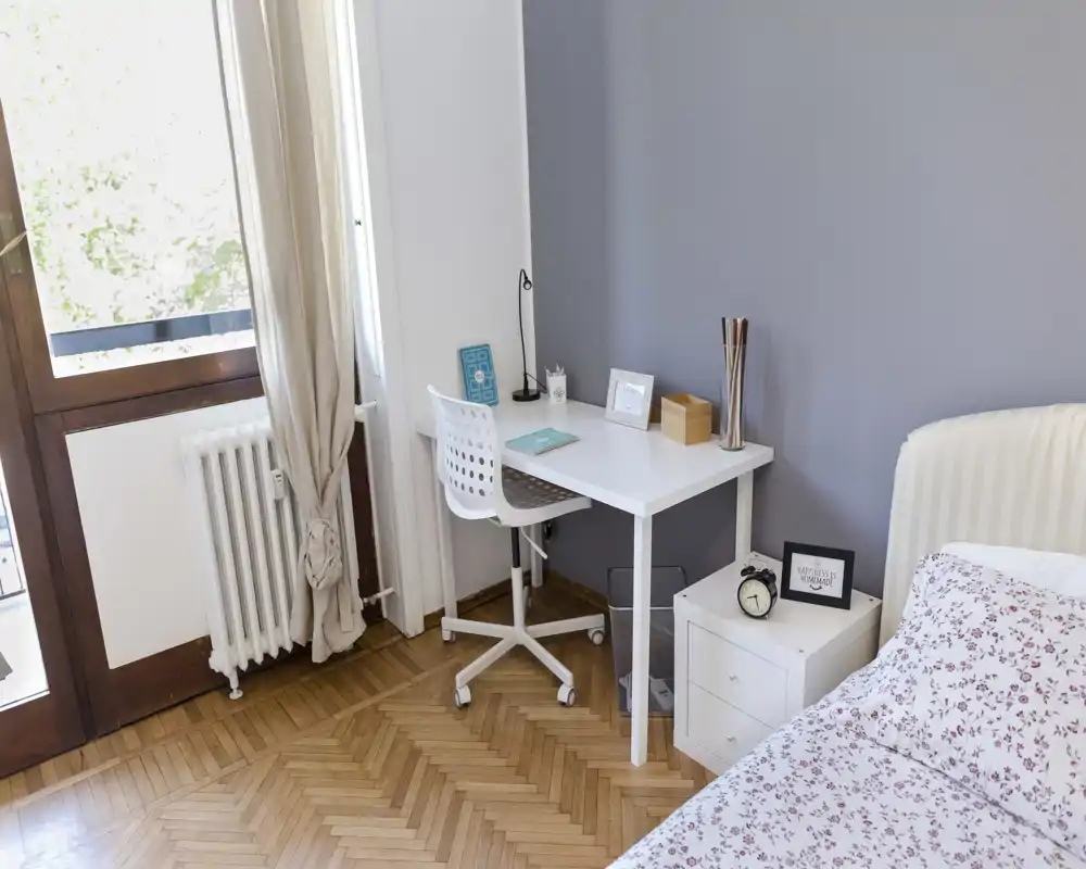 Bright, tidy bedroom with a white bed, floral bedding and a small workspace beside a window. The room features a wooden parquet floor, a bedside table with alarm clock and a compact desk ideal for remote work.