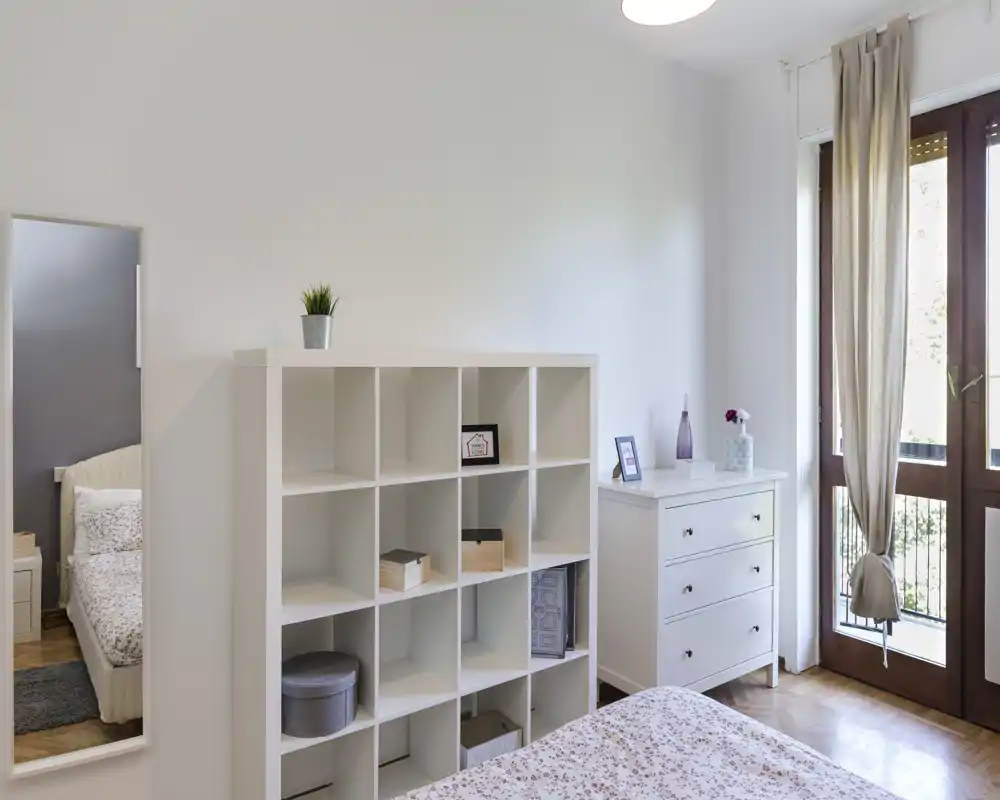 Bright, tidy bedroom with patterned bedding, a storage cube shelf and a dresser beside a large window letting in natural light.