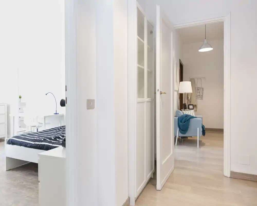Bright, minimal bedroom seen through an open doorway: a white bed with striped bedding and a desk by the window. Clean, naturally lit space ideal for a rental photo.