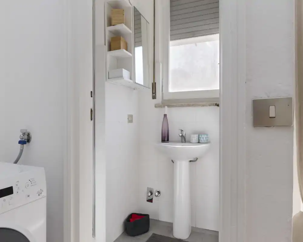 Compact, bright bathroom with a pedestal sink, mirrored cabinet and a nearby washing machine — clean white tiles and natural light from a frosted window.
