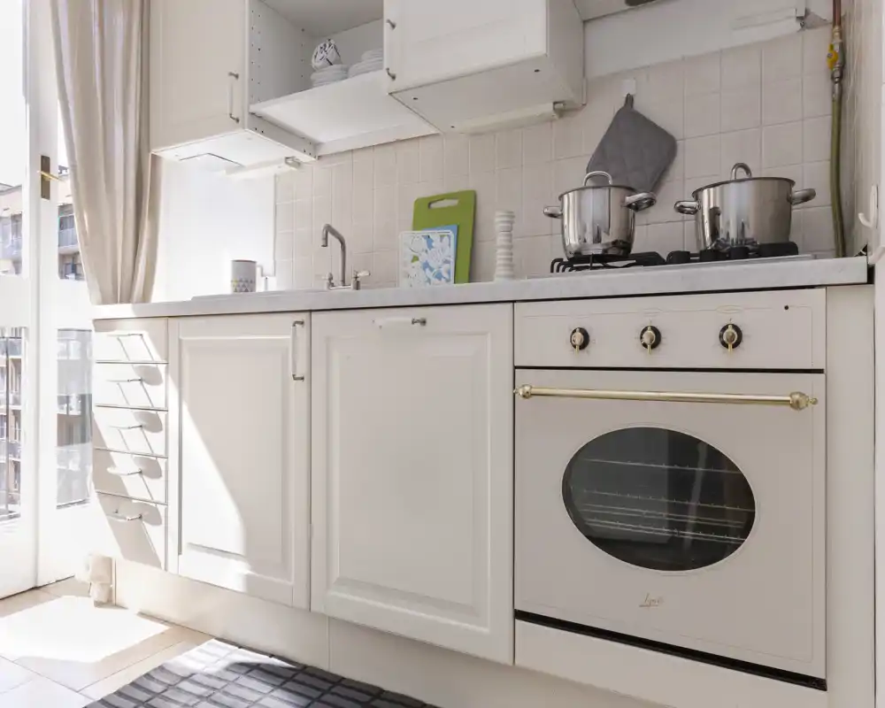 Bright, compact kitchen with a classic white oven, gas hob and a clean countertop bathed in natural light from the window. Thoughtful details like a cutting board, pots and soft curtains create a welcoming, homey atmosphere.