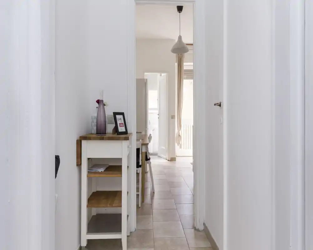 Bright, minimal hallway view leading into a living/dining area with a small wooden console table, pendant light and natural light from the far windows.