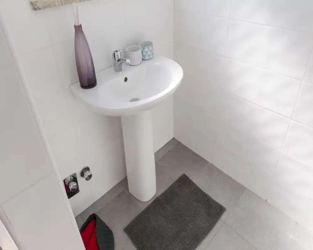 Bright, minimalist bathroom corner with a white pedestal sink, tiled walls and a small floor mat — clean and simple styling ideal for listing photos.