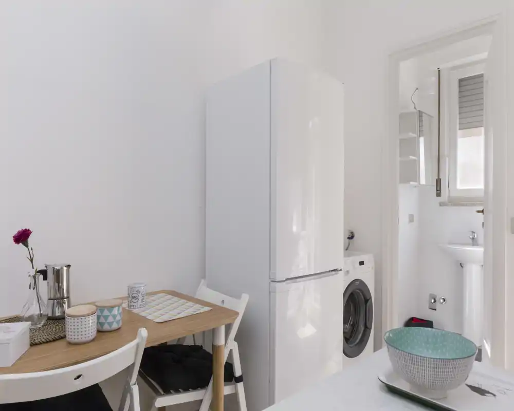 Compact kitchen corner with a small wooden dining table, refrigerator and washing machine; bright, tidy and simply furnished — ideal for a studio or small apartment.