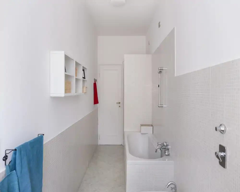 Clean, narrow bathroom with a bathtub on the right, light mosaic tiles halfway up the walls, wall-mounted shelving and bright natural light.