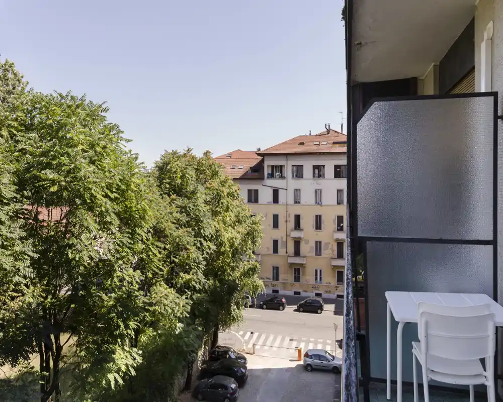 Sunny balcony with a small table and stacked chairs, overlooking leafy trees and a quiet city street with classic residential buildings.