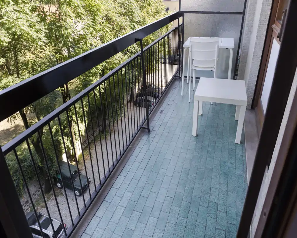 Sunny balcony with turquoise tiled floor, metal railing and a small white table and chairs — an inviting spot for morning coffee overlooking trees.
