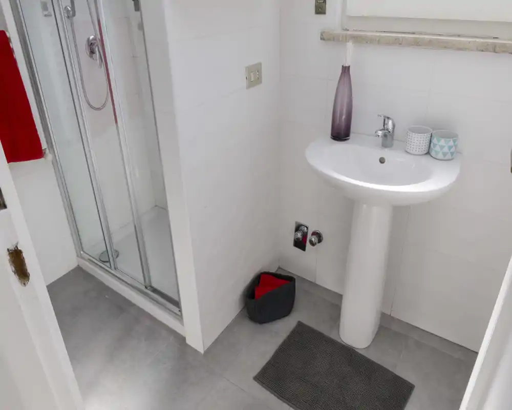 Compact, well-lit bathroom with a pedestal sink, glass shower enclosure and neutral tile flooring — clean and modern.